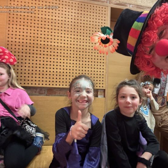 Zwei Mädchen in Kostümen, eines mit Daumen hoch, sitzen neben einem Clown mit roter Nase. Ein Mädchen in Pink steht hinter ihnen, und andere sind anwesend.