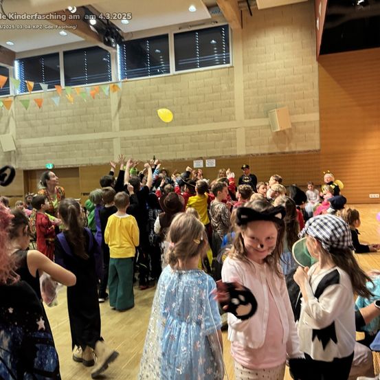 Eine Gruppe von Kindern in Kostümen versammelt sich in einem Saal, wahrscheinlich für ein Fest. Einige halten gelbe Luftballons, und andere tragen Gesichtsbemalung. Ein Mann in einem Kostüm steht in der Mitte.