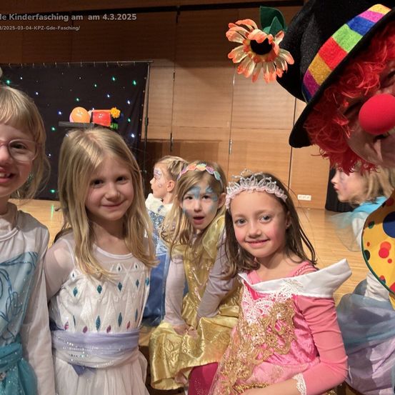 Vier Kinder in Kostümen, eines mit Gesichtsbemalung, posieren für ein Foto mit einem Clown. Sie lächeln und scheinen die Veranstaltung zu genießen. Der Hintergrund zeigt eine Bühne mit schwarzem Hintergrund.