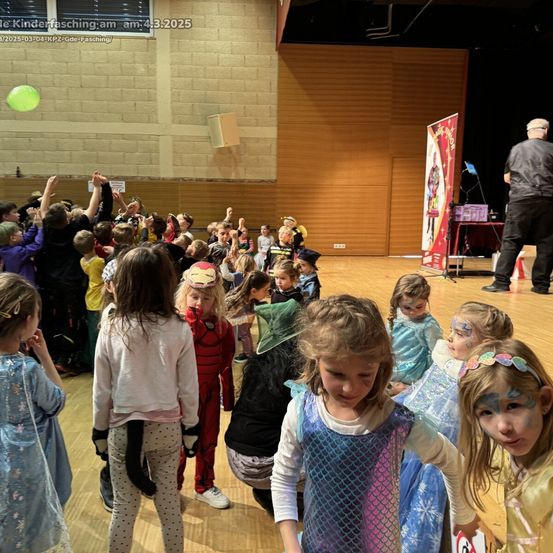 In einem Saal versammeln sich Kinder in Kostümen, einige mit bemalten Gesichtern, ein Mann steht hinter einem Tisch mit einem Schild, ein grüner Ballon schwebt darüber.