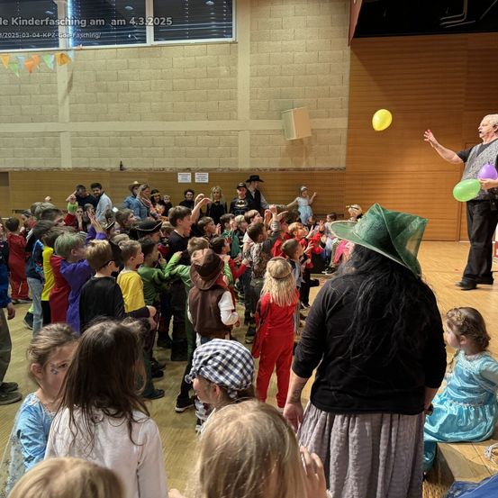 Eine Menge Kinder und Erwachsene versammelt sich in einer Halle für einen Kinderfasching am 4. März 2025. Ein Mann auf der Bühne jongliert mit Luftballons, während eine Frau mit Hut neben ihm steht.