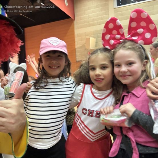 Drei Mädchen, gekleidet in verschiedenen Kostümen, stehen bei einem Kinderkarneval zusammen, lächeln und posieren für ein Foto. Das Mädchen links trägt ein gestreiftes Shirt und hält einen Snack, die mittlere trägt ein Cheerleader-Trikot und das Mädchen rechts trägt ein rosa Shirt mit einer rosa Schleife.