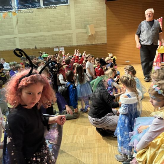 Eine Gruppe von Kindern in verschiedenen Kostümen ist in einem Saal versammelt. Einige sitzen, andere stehen. Ein Erwachsener steht hinten und hält einen gelben Ballon. Das Bild hat einen Wasserzeichen.