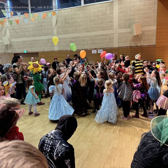 Eine große Gruppe von Kindern in Kostümen, die Luftballons halten, versammelt sich in einem Saal für eine Feier, mit Erwachsenen in der Nähe und bunten Dekorationen an den Wänden.