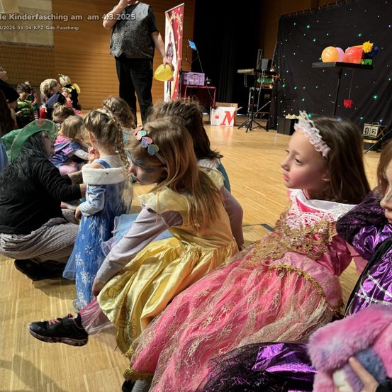 Kinder in Kostümen sehen sich eine Aufführung auf der Bühne an. Ein Mann steht mit Luftballons hinter der Bühne. Einige Kinder sitzen auf dem Boden, während andere hinter ihnen stehen.