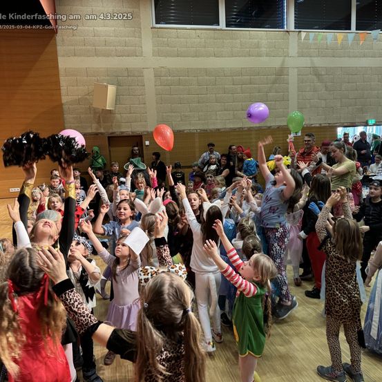 Eine Gruppe von Kindern in Kostümen tanzt und hält Luftballons in einer Halle. Einige heben ihre Hände. Das Datum 4.3.2025 ist oben links sichtbar.