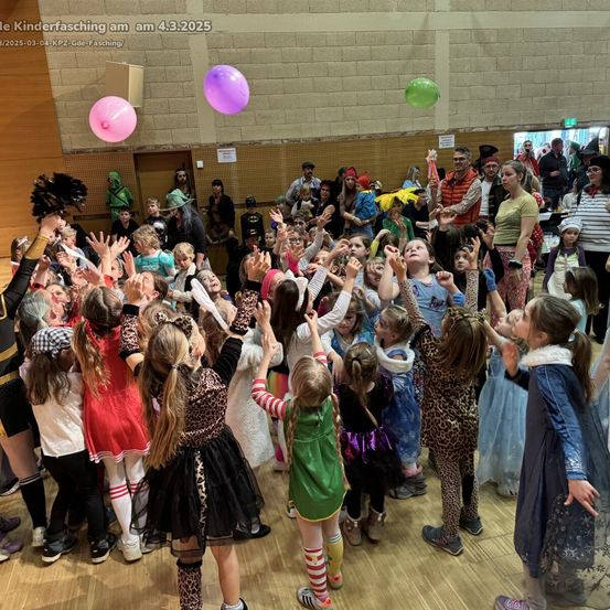 Eine große Menschenmenge aus Kindern und Erwachsenen versammelt sich in einem Innenraum, einige in Kostümen. Sie halten die Hände hoch und einige haben Luftballons. Der Boden ist aus Holz.