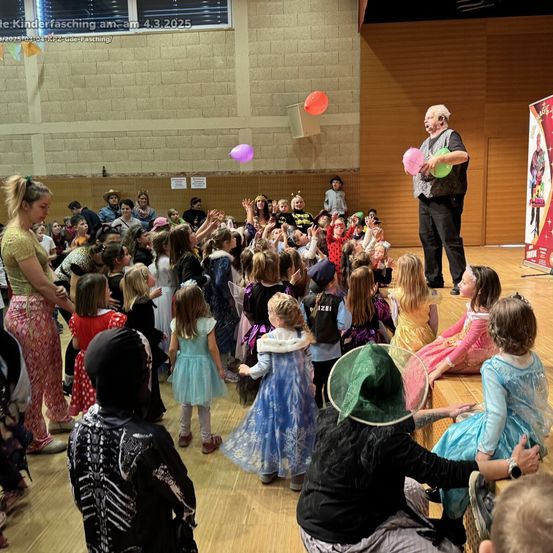 Eine Menschenmenge von Kindern in verschiedenen Kostümen beobachtet einen Mann vor ihnen. Er jongliert mit bunten Ballons. Die Kinder sitzen auf dem Boden. Einige von ihnen tragen Hüte. Ein Banner befindet sich auf der rechten Seite.