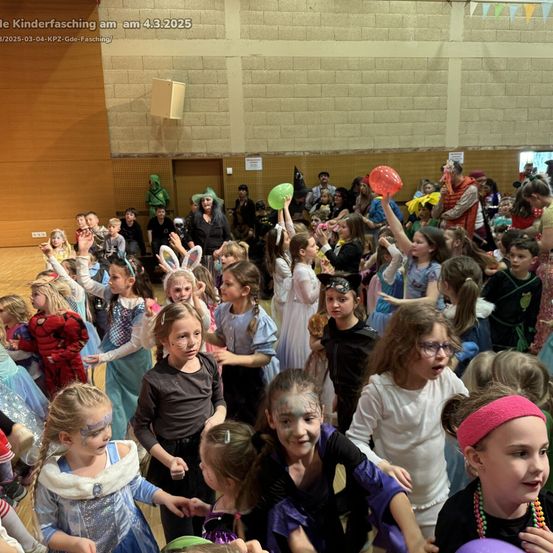 Eine große Gruppe von Kindern in verschiedenen Kostümen, die Luftballons halten, steht in einer Halle. Einige tragen Gesichtsbemalung und halten Plüschtiere. Das Datum, 4.3.2025, ist oben angezeigt.