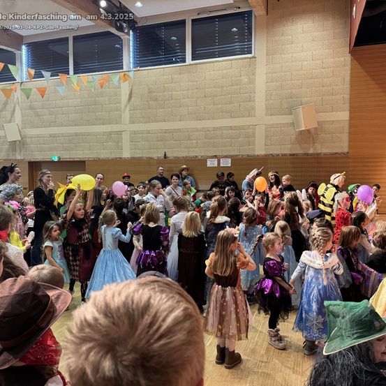 Eine große Gruppe von Kindern in verschiedenen Kostümen, darunter eine Hexe, eine Prinzessin und eine Biene, versammelt sich in einem Saal, einige halten Luftballons, wahrscheinlich für eine Feier.