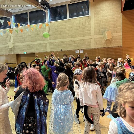 Eine große Gruppe von Kindern in verschiedenen Kostümen tanzt und hebt die Hände in einer Turnhalle. Der Raum ist mit Wimpelketten dekoriert. Ein grüner Ballon schwebt über ihnen.