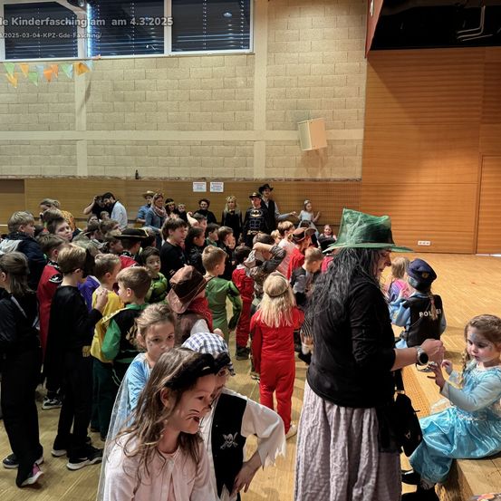Eine Gruppe von Kindern in Kostümen versammelt sich in einem Saal. Eine Erwachsene, die als Hexe verkleidet ist, interagiert mit einem kleinen Mädchen in einem blauen Kleid. Das Bild stammt vom Kinderfasching am 4. März 2025.