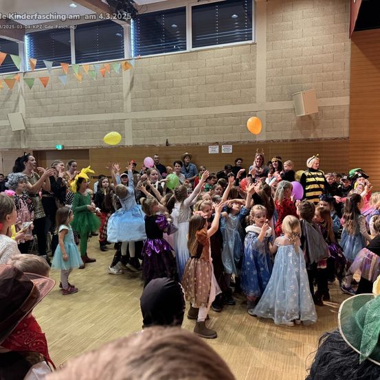 Eine Gruppe von Kindern in Kostümen und Masken ist in einem Saal versammelt, einige heben ihre Hände, und ein paar Ballons schweben in der Luft.