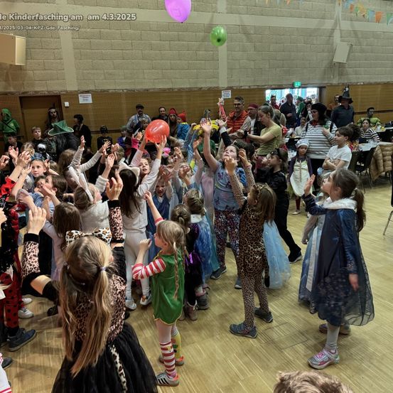 Kinder in verschiedenen Kostümen tanzen in einem großen Raum und halten Luftballons. Eine Frau blickt von hinten zu, andere beobachten von den Seiten. Ein grüner Ballon schwebt oben.
