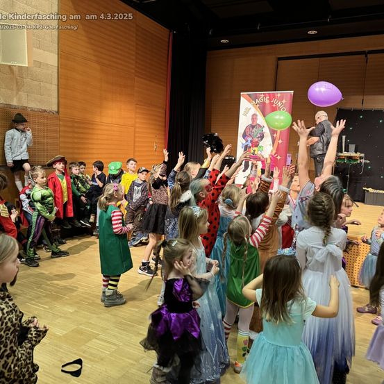 Eine Gruppe von Kindern in verschiedenen Kostümen ist auf einer Bühne versammelt und hebt die Hände. Einige tragen Superheldenkostüme, während andere als Hexen oder Feen gekleidet sind. Hinter ihnen ist ein Banner mit dem Text 'Magic Juno' zu sehen.