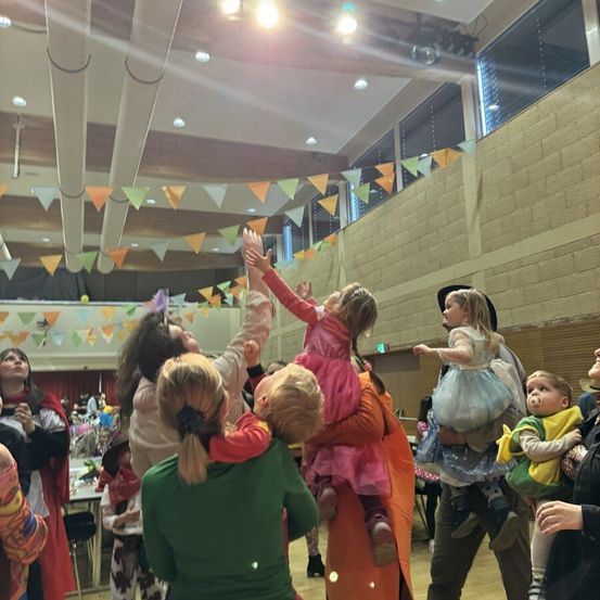 Eine Gruppe von Erwachsenen und Kindern befindet sich in einer Halle und hebt die Hände, wahrscheinlich für ein Foto. Bunte Flaggen hängen von der Decke.
