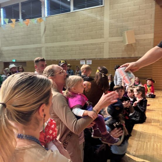 Eine Gruppe von Menschen, einschließlich Erwachsener und Kinder, ist drinnen versammelt. Einige klatschen, während andere Kinder halten. Eine Frau hält ein Kleinkind, während ein Mann ein Baby hält. Eine Hand ist sichtbar und hält ein Buch.