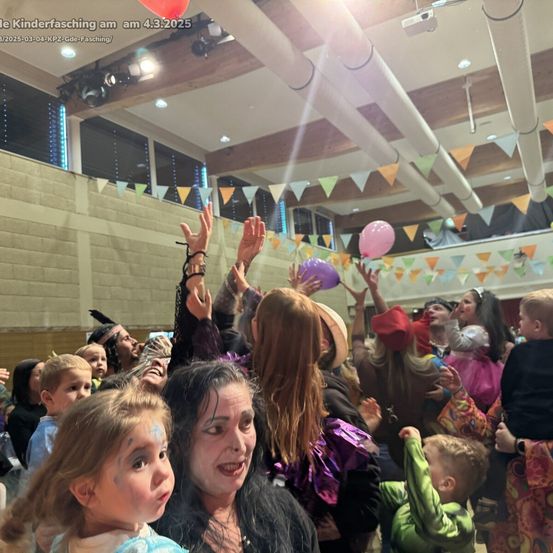 Eine Menschenmenge, bestehend aus Kindern und Erwachsenen, feiert einen Karneval in einem dekorierten Raum mit bunten Bannern und Luftballons.