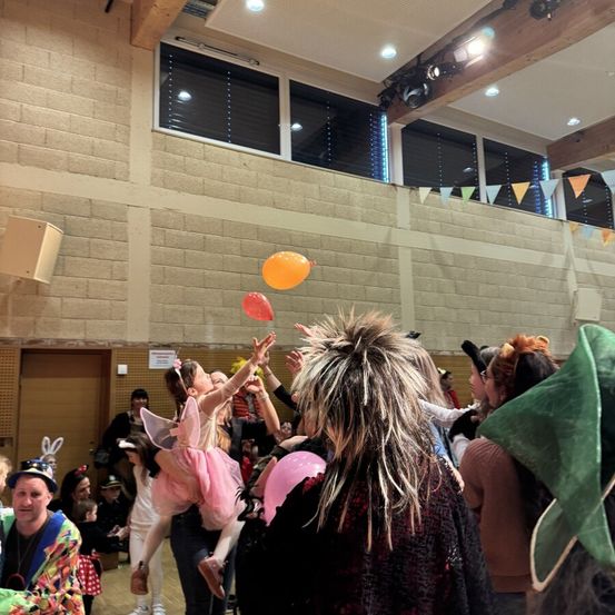 Eine große Gruppe von Menschen in einer Halle, viele in Kostümen gekleidet. Ein Mädchen in einem Feenkostüm streckt sich nach einem Ballon.