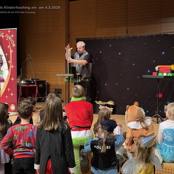 Ein Mann führt eine Zaubershow auf der Bühne für eine Gruppe von Kindern in Kostümen vor. Die Bühne ist mit einem schwarzen Hintergrund und einem roten Banner dekoriert.