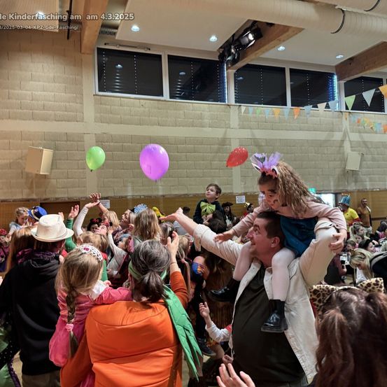 Eine Menschenmenge versammelt sich in einer Halle, einige heben ihre Hände, und ein Mann trägt ein Kind auf seinen Schultern. Es gibt Luftballons und Wimpel, die von der Decke hängen. Das Bild hat einen Wasserzeichen, der 'Kinderfasching am 4.3.2025' lautet.