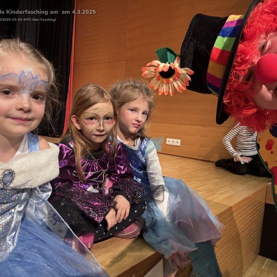 Drei junge Mädchen in Kostümen sitzen zusammen auf einer Bühne. Sie haben Gesichtsbemalung. Dahinter steht ein Clown mit einer Blume und eine Figur in Streifen. Das Bild ist vom Kinderfasching-Event am 4. März 2025.