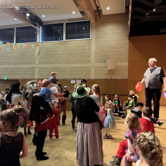 Eine Menschenmenge, darunter Kinder in Kostümen, versammelte sich am 4.3.2025 in einem Saal für Kinderfasching. Ein Mann steht auf der Bühne, hält Luftballons, und eine Frau mit einem Kind ist anwesend.