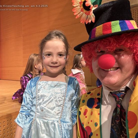 Ein junges Mädchen in einem blauen Kleid und ein Mann in einem Clownskostüm posieren zusammen und lächeln in die Kamera. Hinter ihnen stehen einige Kinder in Kostümen auf der Bühne.