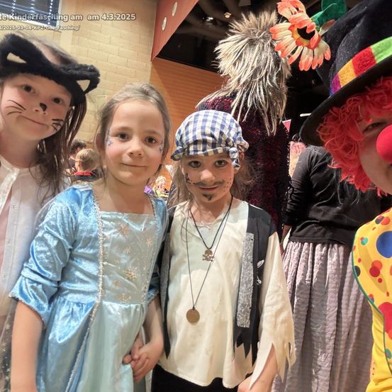 Vier Kinder in Kostümen, zwei mit Gesichtsbemalung, stehen zusammen. Ein Mädchen in einem blauen Kleid und ein Junge in einem Piratenhut. Dahinter stehen Erwachsene in Kostümen.