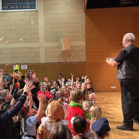 Eine Gruppe von Kindern und Erwachsenen ist in einem Raum versammelt. Ein Mann mit Mikrofon steht vor ihnen. Einige Kinder tragen Kostüme. Konfetti ist in der Luft. Eine digitale Uhr zeigt die Zeit als 4:30.