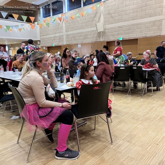 Eine große Gruppe von Menschen, Erwachsenen und Kindern, sitzt um Tische herum in einem Saal. Einige tragen Kostüme. Eine Frau mit rosa Leggings und ein Mädchen mit Mickey-Mouse-Ohren sitzen im Vordergrund.