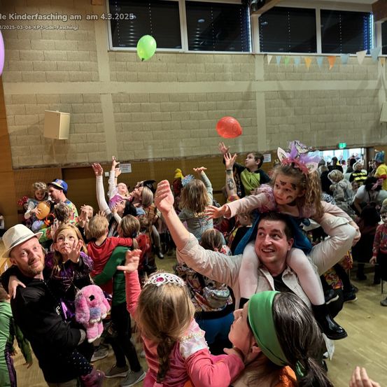 Eine große Gruppe von Menschen ist drinnen während eines Karnevals oder einer Party. Viele Kinder sind in Kostümen gekleidet und heben ihre Hände. Zwei Erwachsene tragen Kinder auf ihren Schultern. Luftballons schweben in der Luft.