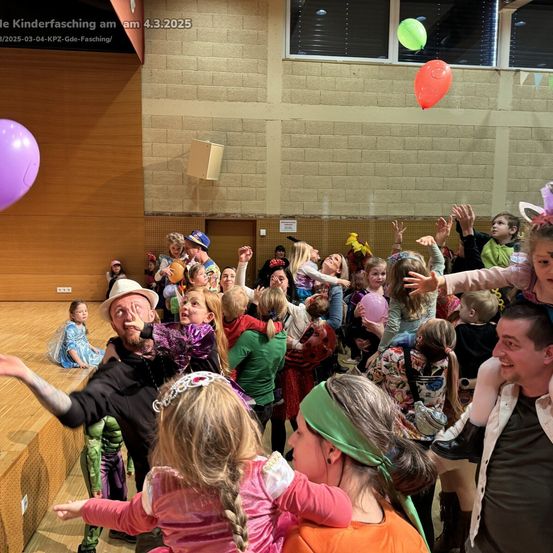 Eine Gruppe von Menschen in Kostümen versammelt sich drinnen, mit Kindern, die herumspielen, und schwebenden Luftballons. Ein Mann in der Mitte hält ein Kind auf seinen Schultern.