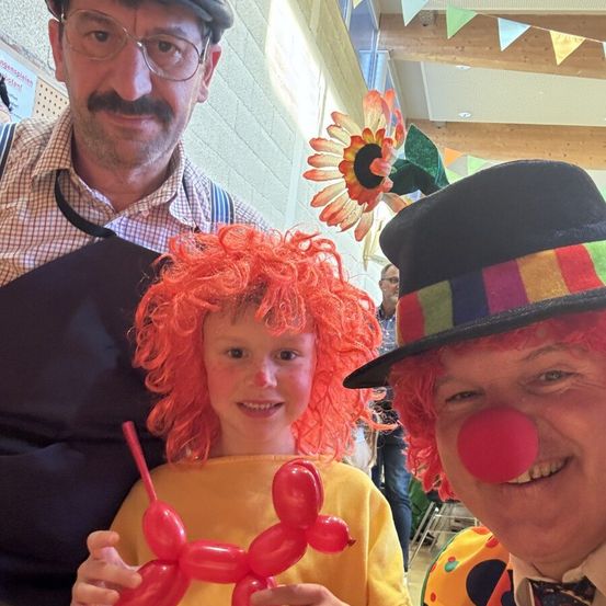 Drei Personen stehen zusammen; eine hält ein Ballontier, eine andere trägt eine Clownsnase und die dritte ist in einem karierten Hemd gekleidet. Sie scheinen für ein Foto zu posieren.