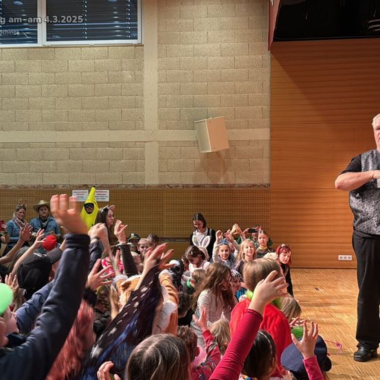 Ein Mann steht auf der Bühne, während viele Kinder vor ihm jubeln. Die Kinder heben ihre Hände und einige halten Luftballons. Der Mann hält ein Mikrofon. Das Bild hat einen Zeitstempel vom 4.3.2025.