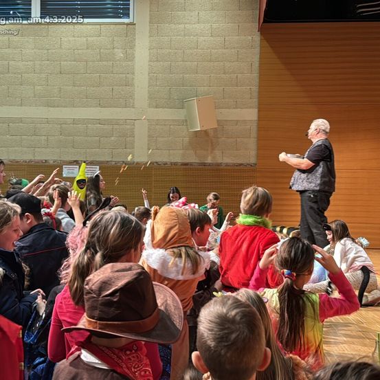 Ein Mann steht auf einer Bühne vor einer Gruppe von Kindern. Einige Kinder tragen Kostüme. Der Raum hat Ziegelwände und eine Holzbühne.