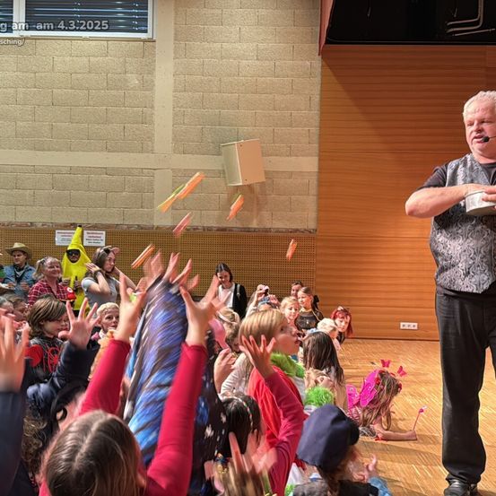 Ein Mann spricht in ein Mikrofon, während mehrere Kinder die Hände heben und ihn ansehen. Es gibt Objekte, die in der Luft fliegen.