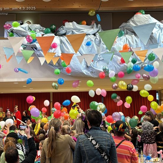 Eine große Menschenmenge in einem Raum voller bunter Luftballons und Wimpel, die von der Decke hängen. Die Menschen halten Ballons hoch und einige tragen Kostüme.