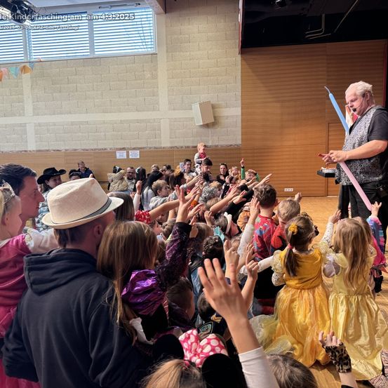 Ein Mann mit Mikrofon unterhält eine Menge von Kindern in Kostümen, viele heben ihre Hände in Begeisterung. Der Ort scheint ein Saal zu sein, mit Holzwänden und einem Fenster im Hintergrund.