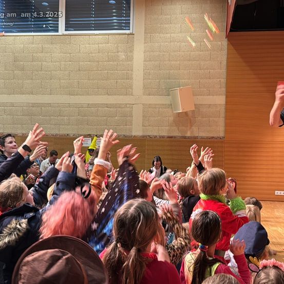 Eine Menschenmenge aus Kindern und Erwachsenen in einem Saal, die ihre Hände heben, einige tragen Hüte und bunte Kostüme. Ein Erwachsener steht vor ihnen, möglicherweise bei einer Aufführung. Die Wand dahinter hat ein Ziegelmuster und ein Fenster.