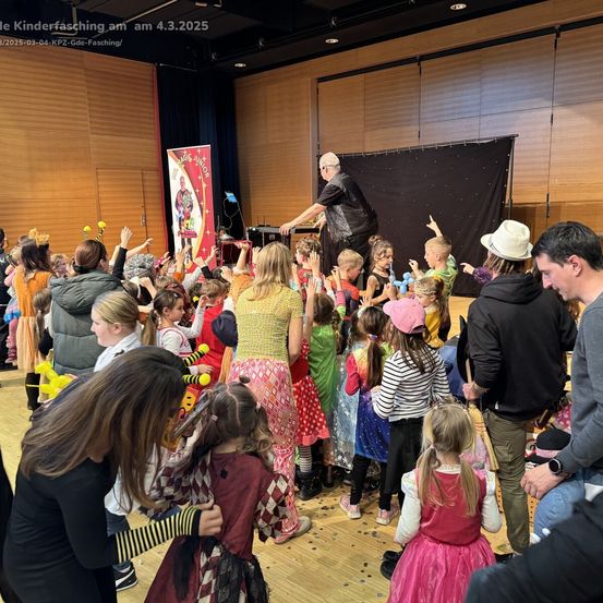 Eine Gruppe von Kindern in Kostümen umgibt einen Mann, der auf einer Bühne Zaubertricks vorführt. Die Veranstaltung ist ein Kinderfasching am 4. März 2025.
