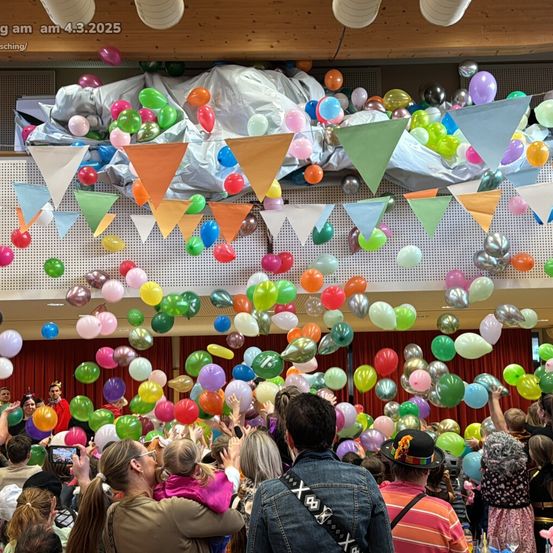 Eine Menschenmenge in einem Saal, einige halten Luftballons, andere schauen mit bunten Dreiecksfahnen an der Decke auf, wo bunte Luftballons schweben.