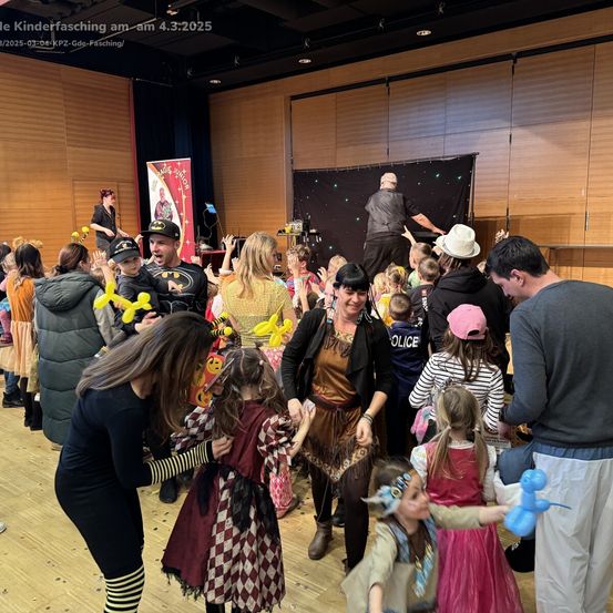 Eine Gruppe von Menschen, darunter Erwachsene und Kinder, versammelt sich in einem Raum, möglicherweise für einen Kinderfasching. Einige tragen Kostüme, und eine Frau hält einen Luftballon. Ein Mann steht hinter einer Bühne mit einem Banner.