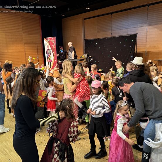 Menge von Kindern in Kostümen bei einem Karneval, versammelt um eine Bühne, auf der ein Zauberer auftritt, mit Erwachsenen, die beaufsichtigen, und einem Hintergrund von schwarzen und weißen Lichtern.