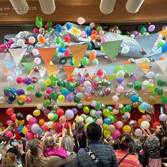 Eine große Menschenmenge, darunter Erwachsene und Kinder, versammelt sich in einem Raum voller bunter Luftballons, von denen einige schweben. Sie heben die Hände und genießen die festliche Atmosphäre. Der Raum hat eine Holzdecke mit hängenden Dekorationen und einem roten Vorhang im Hintergrund.