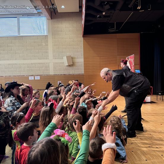 Ein Mann führt einen Zaubertrick in einem großen Saal vor, umgeben von Kindern, die ihre Hände heben. Die Bühne ist aus Holz mit einem Banner und einem schwarzen Vorhang im Hintergrund.
