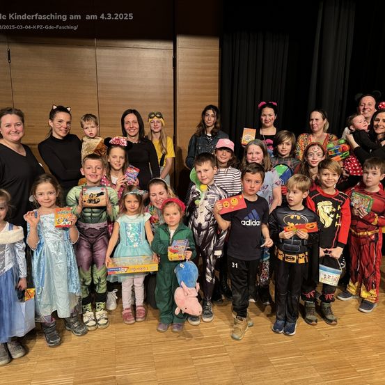 Gruppenfoto von Kindern in Kostümen bei einer Karnevalsveranstaltung am 4. März 2025. Einige halten Geschenke, andere tragen Superheldenkostüme, und der Hintergrund zeigt eine Holzwand und einen schwarzen Vorhang.
