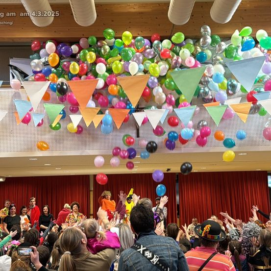 Eine lebhafte Indoor-Party mit einer Menge Menschen in festlicher Kleidung, umgeben von bunten Luftballons und Bannern, die von der Decke hängen.