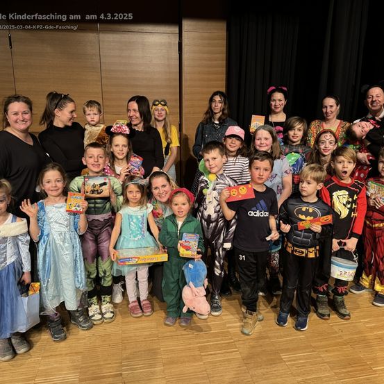 Eine Gruppe von Kindern und Erwachsenen in Kostümen posiert für ein Foto auf der Bühne, hält Geschenke und Spielzeug. Das Datum 4.3.2025 ist oben sichtbar.
