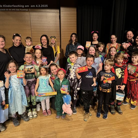 Eine Gruppe von Kindern und Erwachsenen in Kostümen posiert für ein Foto auf einem Holzboden. Sie halten Geschenke und haben Gesichtsbemalung. Der Hintergrund besteht aus einer Holzwand und schwarzen Vorhängen. Das Datum ist der 4. März 2025.